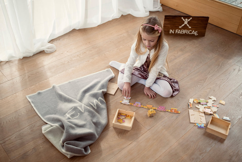 Bambina che gioca con i giocattoli, accanto a una copertina personalizzata in cashmere. Comfort e divertimento in un ambiente accogliente, con dettagli artigianali.