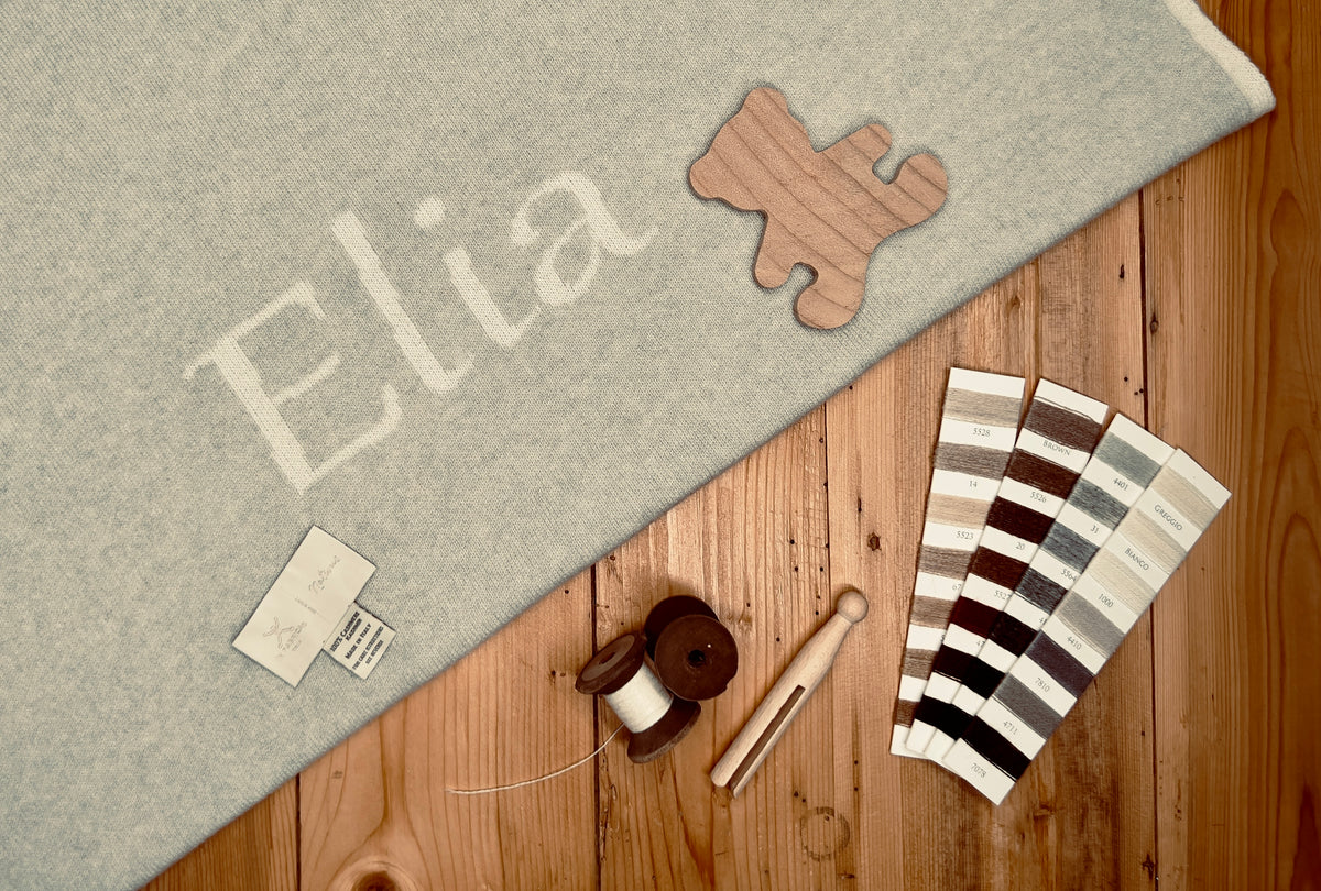 Copertina personalizzata con nome su un tavolo di legno, affiancata da cartelle colori e un orsetto in legno di cedro. Eleganza e personalizzazione in prodotti artigianali per bambini.